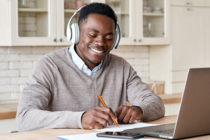 Young man smiling while wearing headphones and taking notes in front of a laptop
